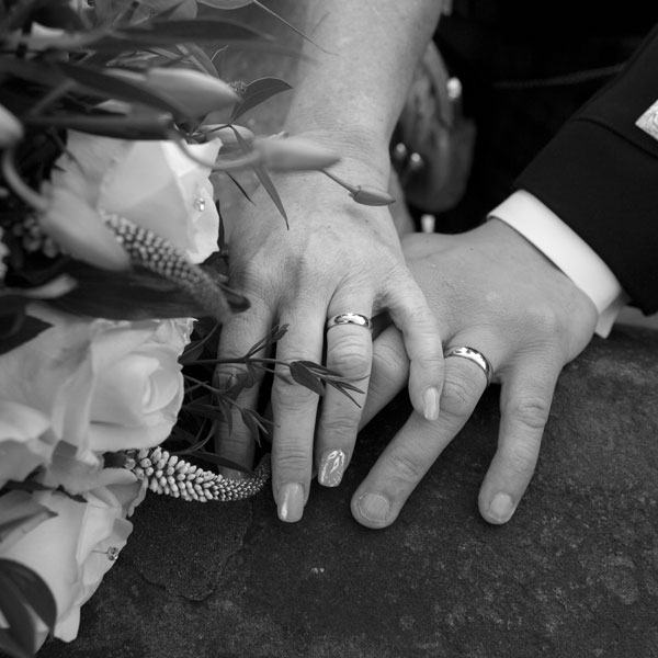 Bride and groom hands on top of one another showing their wedding bands next to the bouquet on a stone wall in black and white