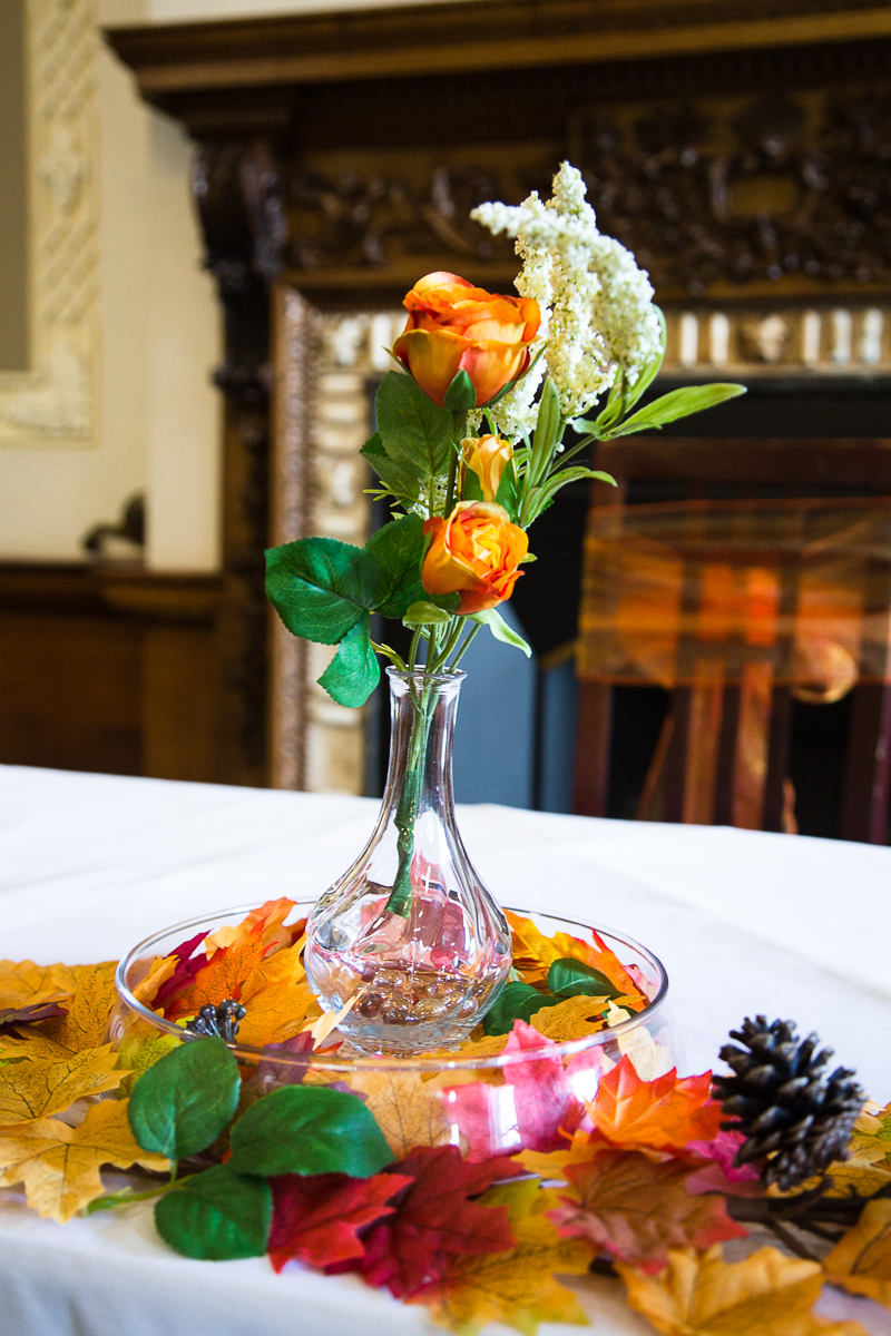 Autumn theme centrepiece with two orange flowers in a small glass vase surrounded by autumn orange yellow and green leaves